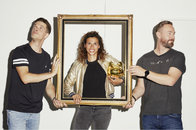 A woman with a piggy bank, standing behind a frame held by two men.